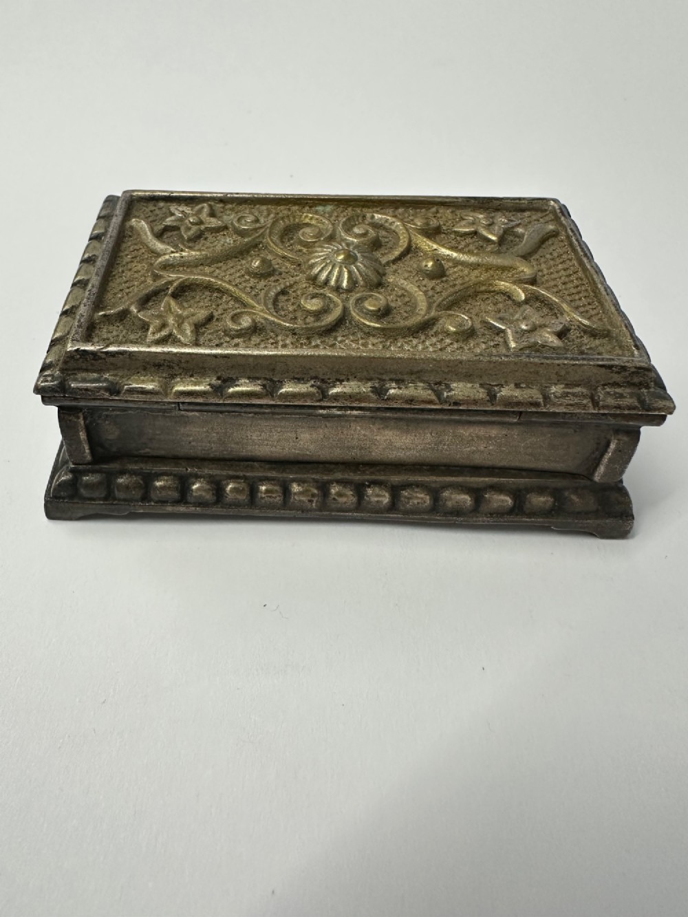An antique silvered brass desktop vesta box with a hinged floral embossed lid, ribbed striker strip, and front tray resting on a beaded base. View III