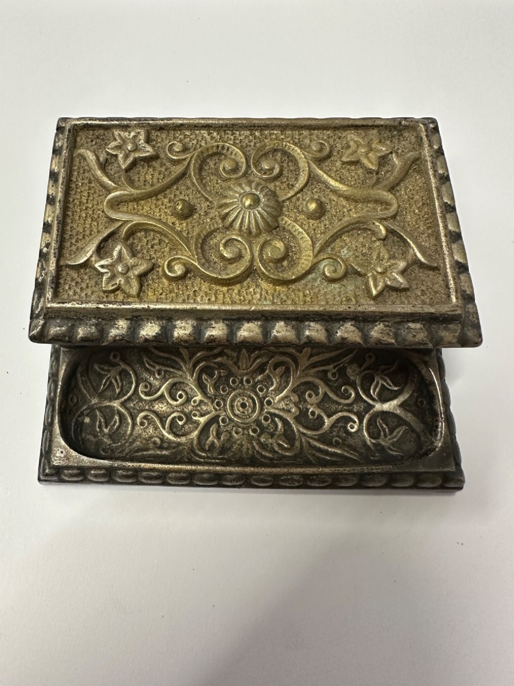 An antique silvered brass desktop vesta box with a hinged floral embossed lid, ribbed striker strip, and front tray resting on a beaded base. View IV