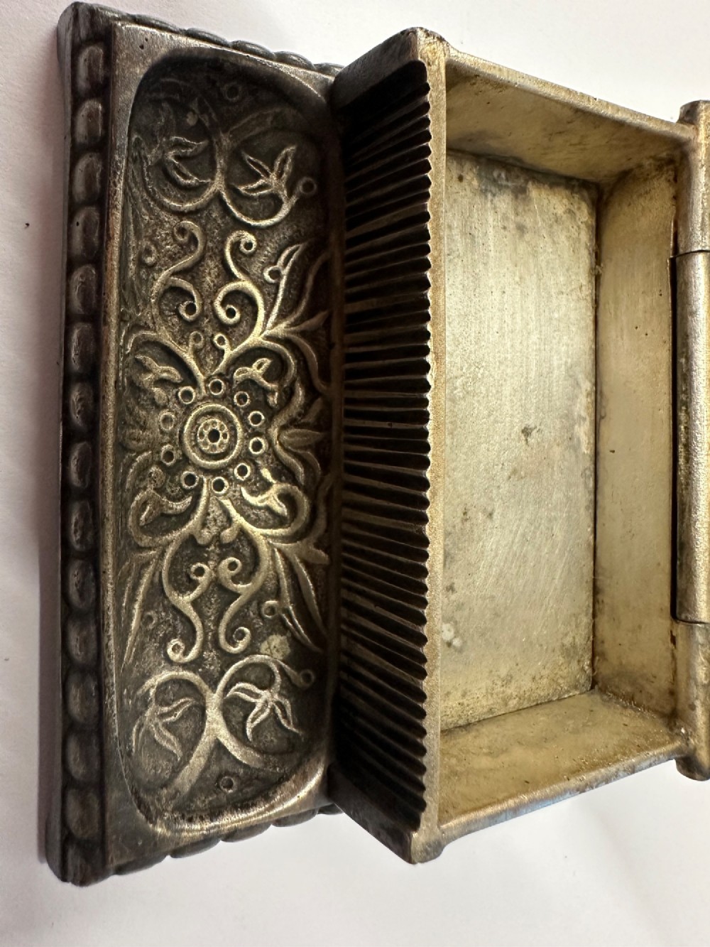 An antique silvered brass desktop vesta box with a hinged floral embossed lid, ribbed striker strip, and front tray resting on a beaded base. View V