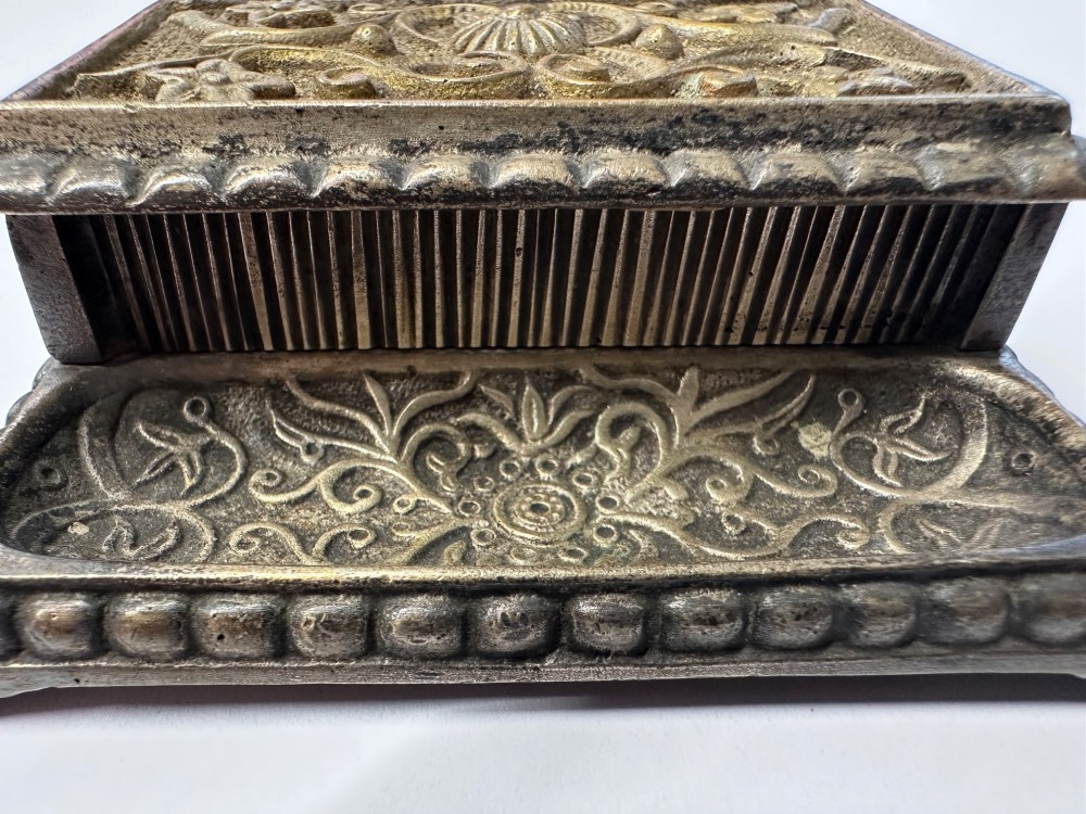 An antique silvered brass desktop vesta box with a hinged floral embossed lid, ribbed striker strip, and front tray resting on a beaded base. View VI
