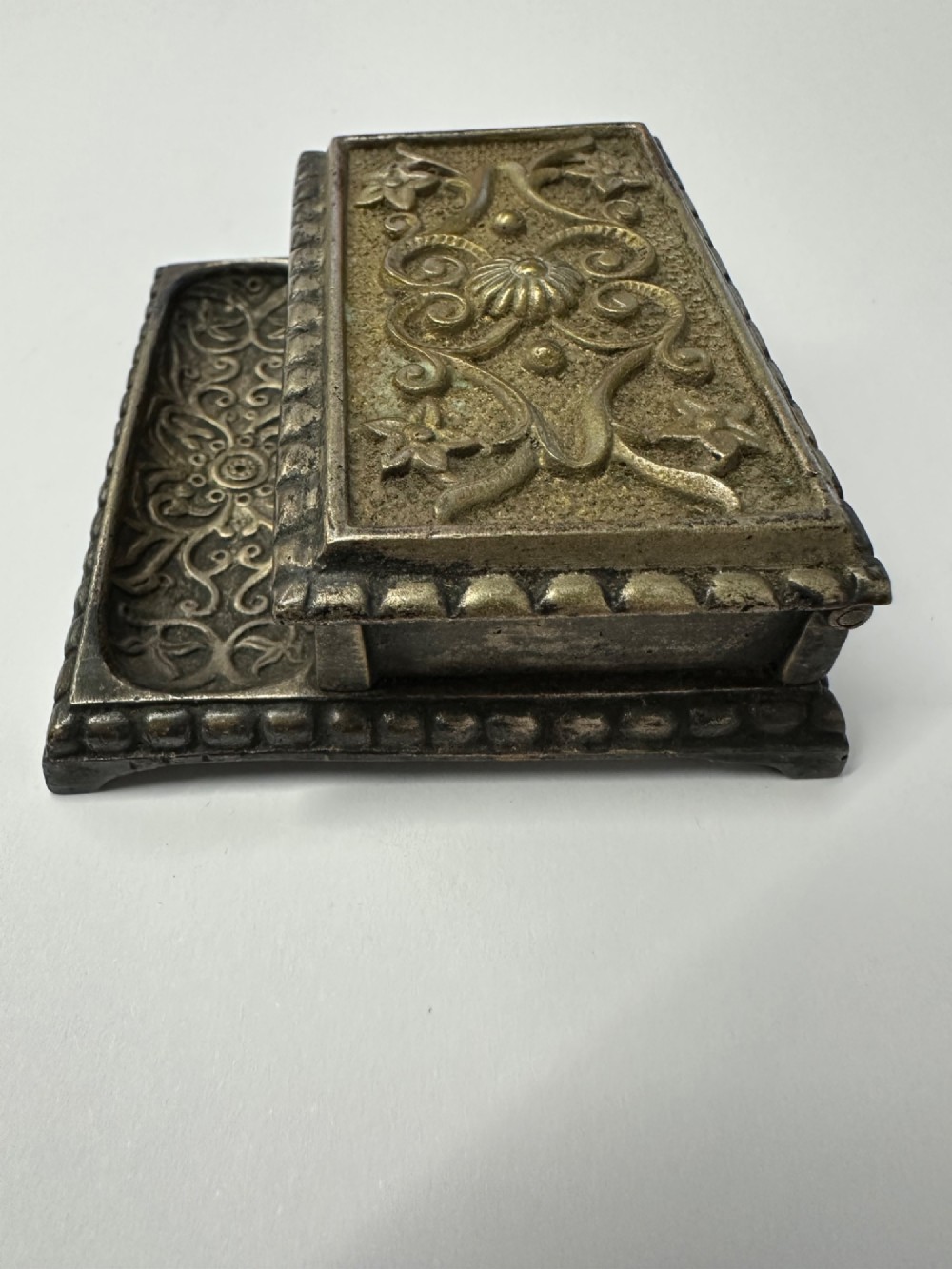 An antique silvered brass desktop vesta box with a hinged floral embossed lid, ribbed striker strip, and front tray resting on a beaded base. View IX
