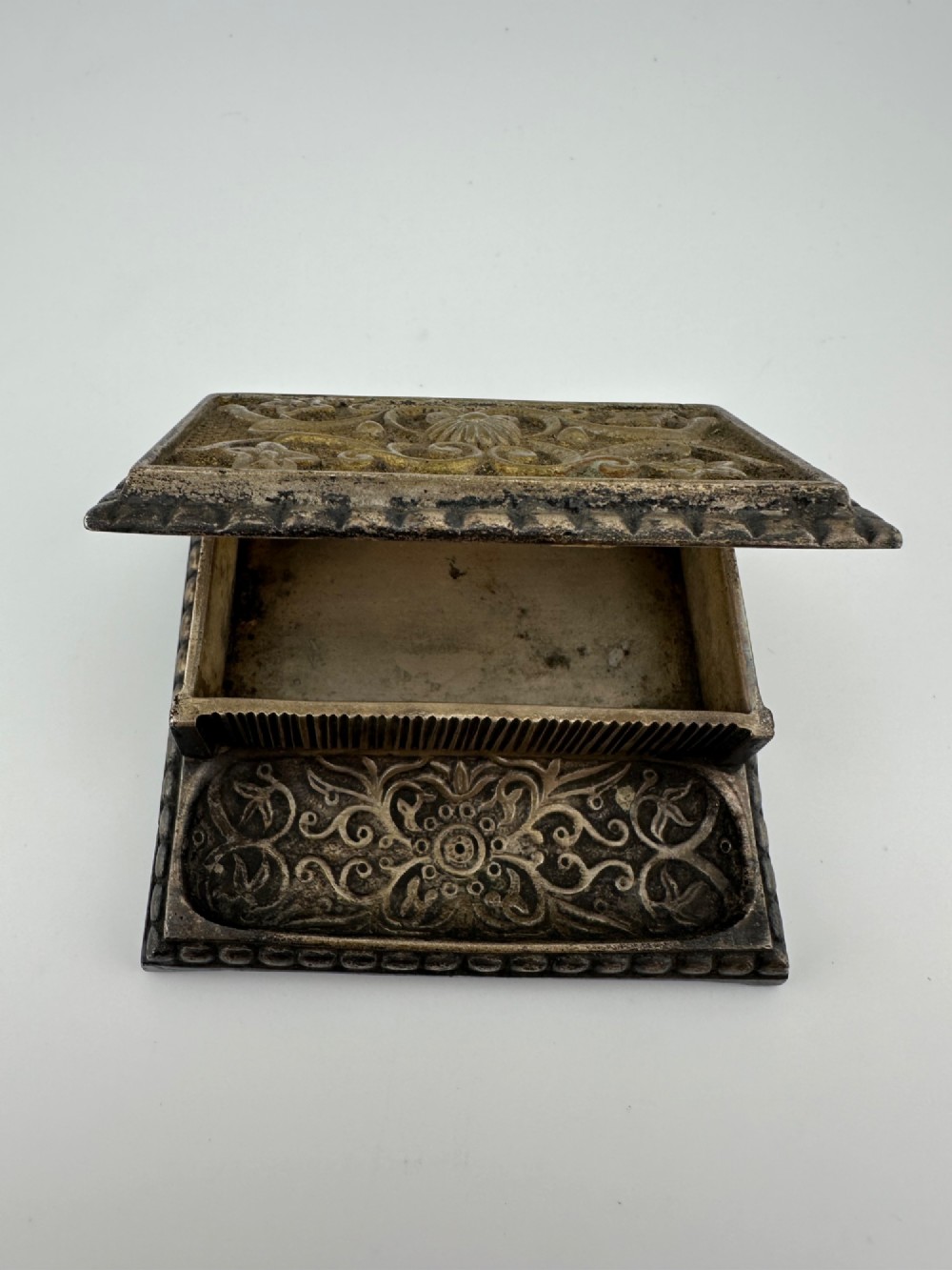 An antique silvered brass desktop vesta box with a hinged floral embossed lid, ribbed striker strip, and front tray resting on a beaded base. View XI