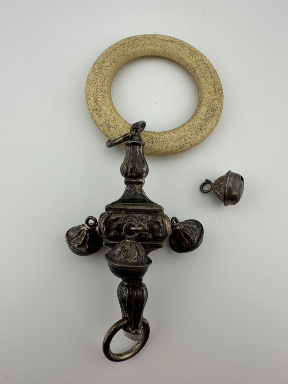 An antique silver baby rattle featuring embossed metalwork, small attached bells, and a cream-coloured circular teether ring.