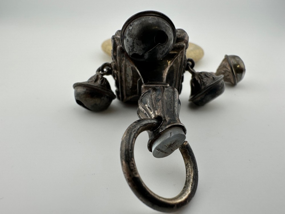 An antique silver baby rattle featuring embossed metalwork, small attached bells, and a cream-coloured circular teether ring. View II