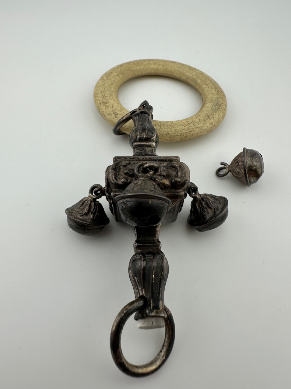 An antique silver baby rattle featuring embossed metalwork, small attached bells, and a cream-coloured circular teether ring. View III