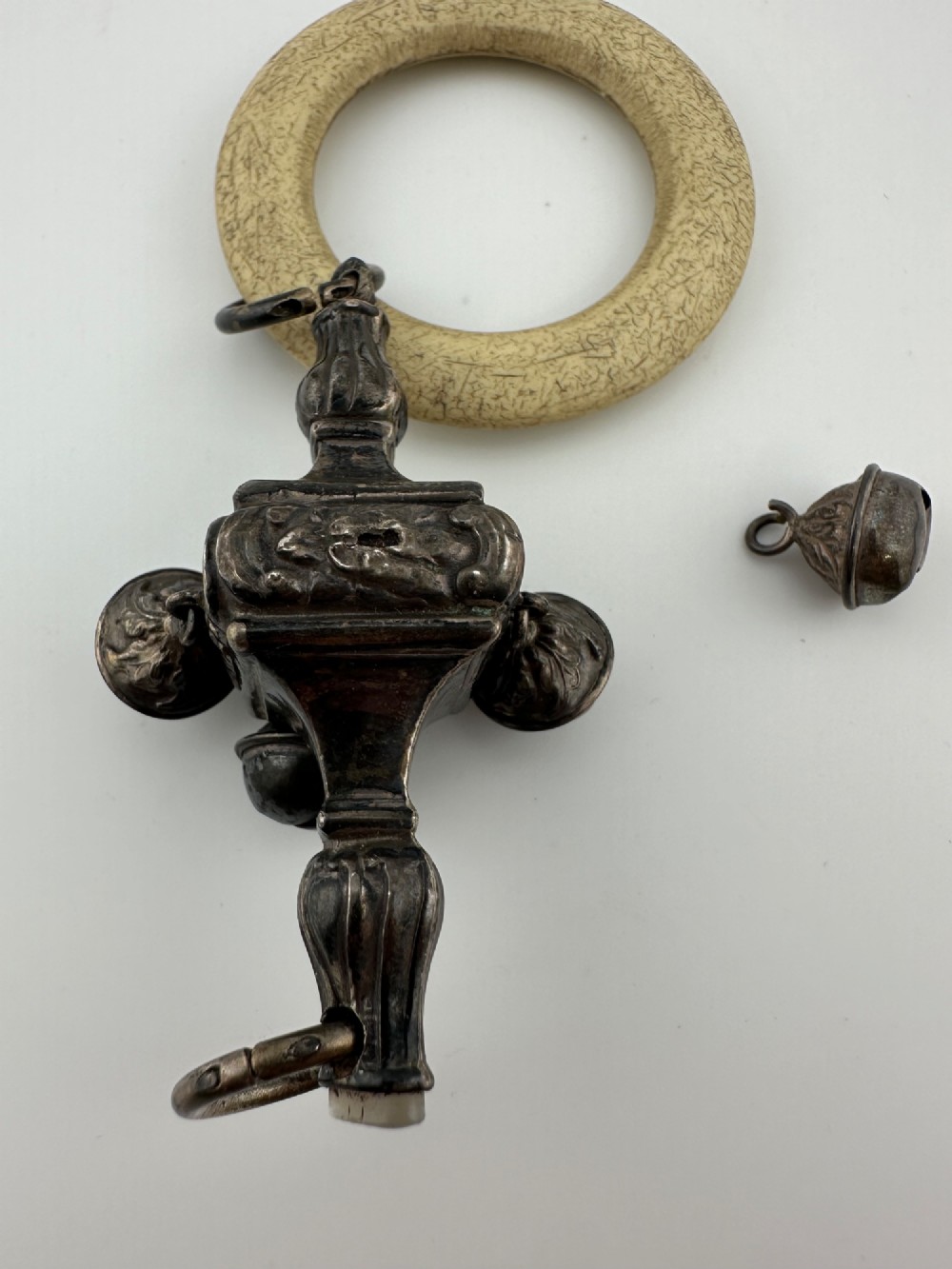 An antique silver baby rattle featuring embossed metalwork, small attached bells, and a cream-coloured circular teether ring. View IV