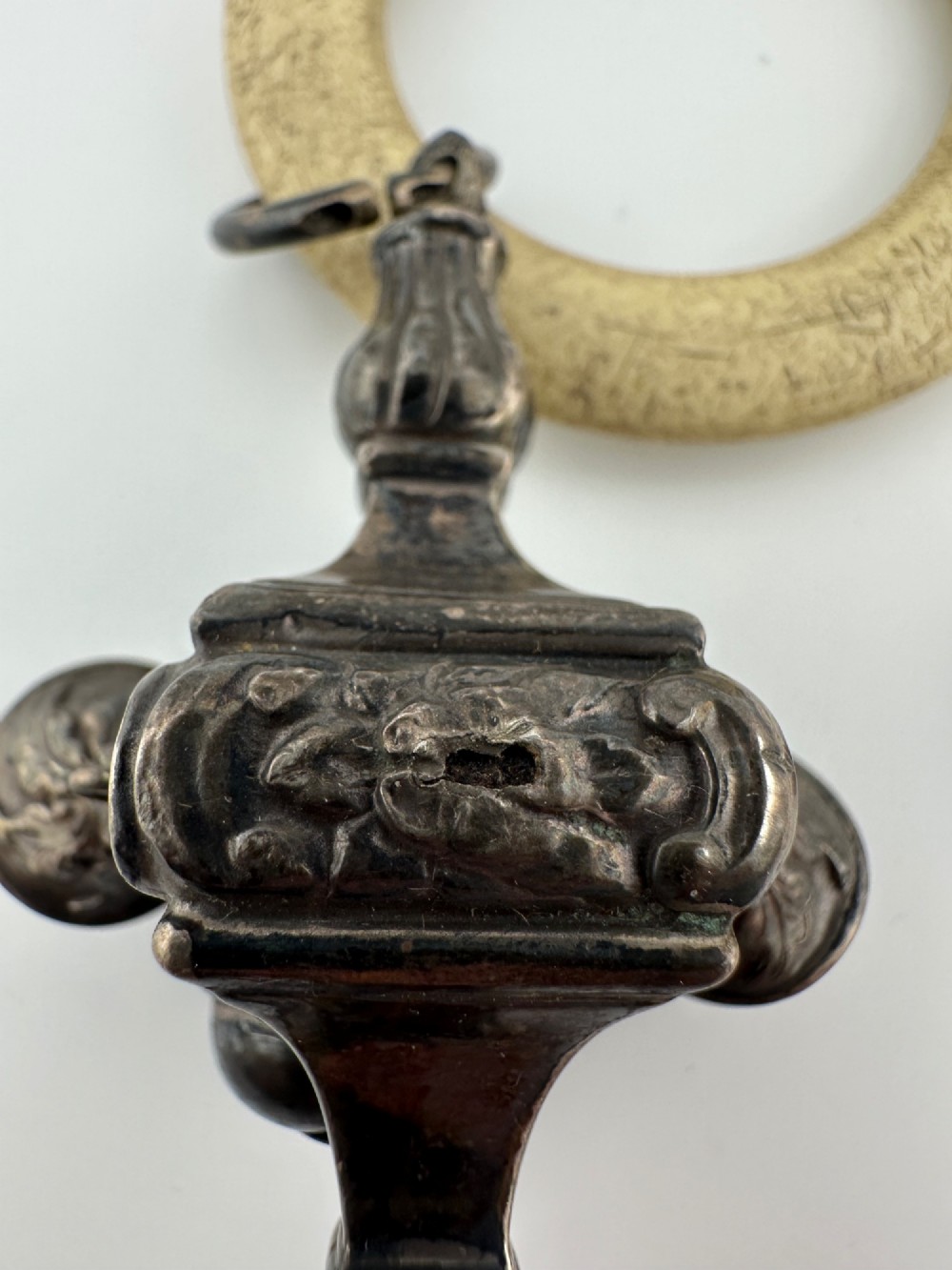 An antique silver baby rattle featuring embossed metalwork, small attached bells, and a cream-coloured circular teether ring. View V