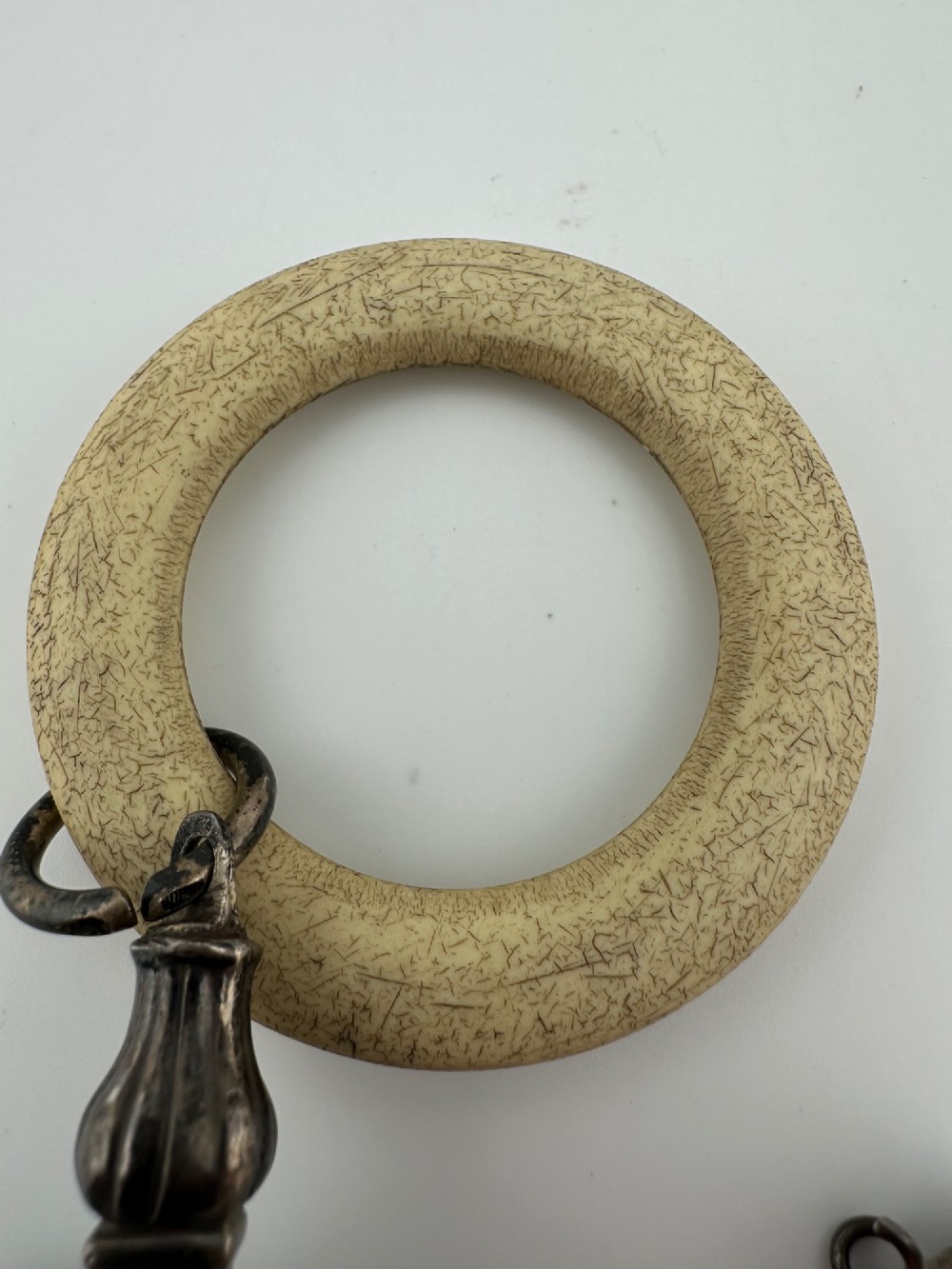An antique silver baby rattle featuring embossed metalwork, small attached bells, and a cream-coloured circular teether ring. View VI