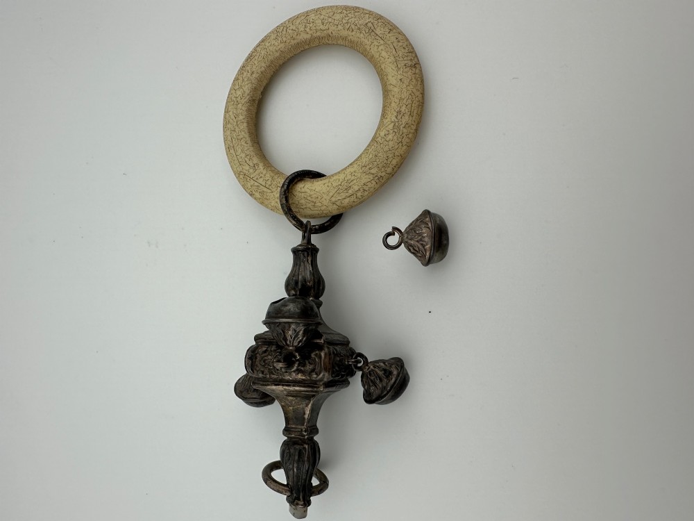 An antique silver baby rattle featuring embossed metalwork, small attached bells, and a cream-coloured circular teether ring. View VII