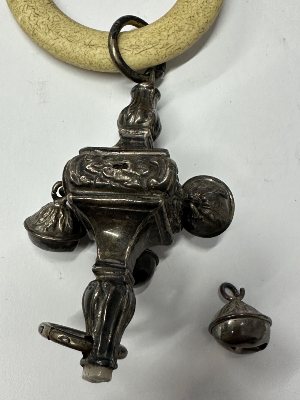 An antique silver baby rattle featuring embossed metalwork, small attached bells, and a cream-coloured circular teether ring. View X