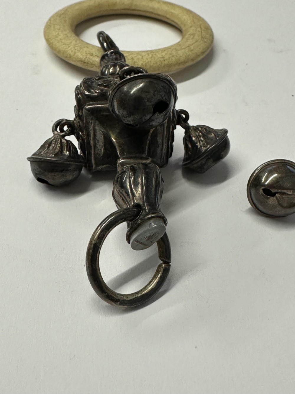 An antique silver baby rattle featuring embossed metalwork, small attached bells, and a cream-coloured circular teether ring. View XI