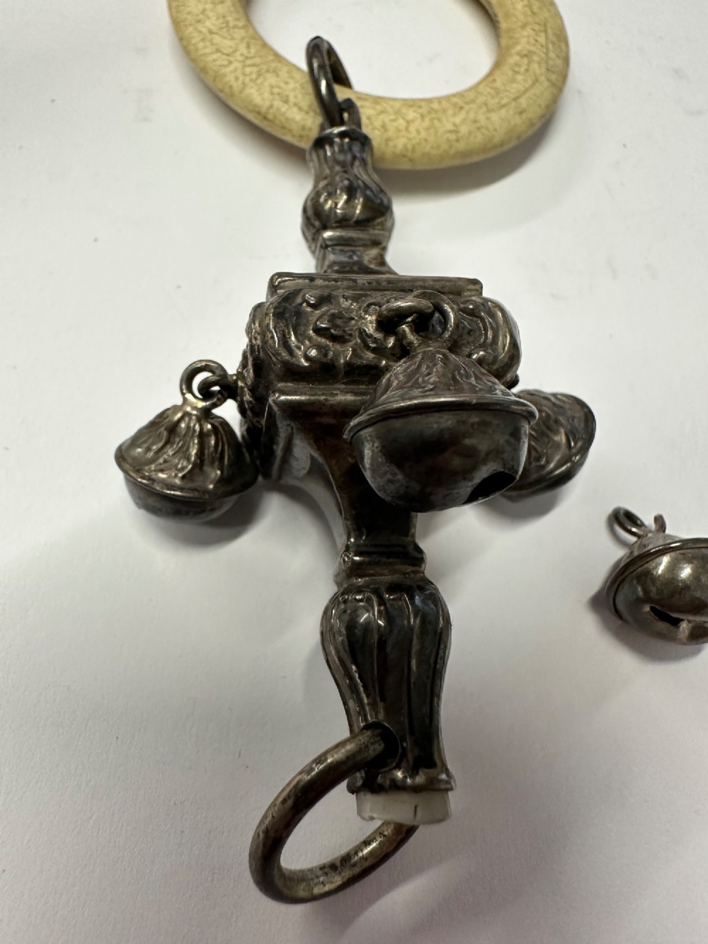 An antique silver baby rattle featuring embossed metalwork, small attached bells, and a cream-coloured circular teether ring. View XII
