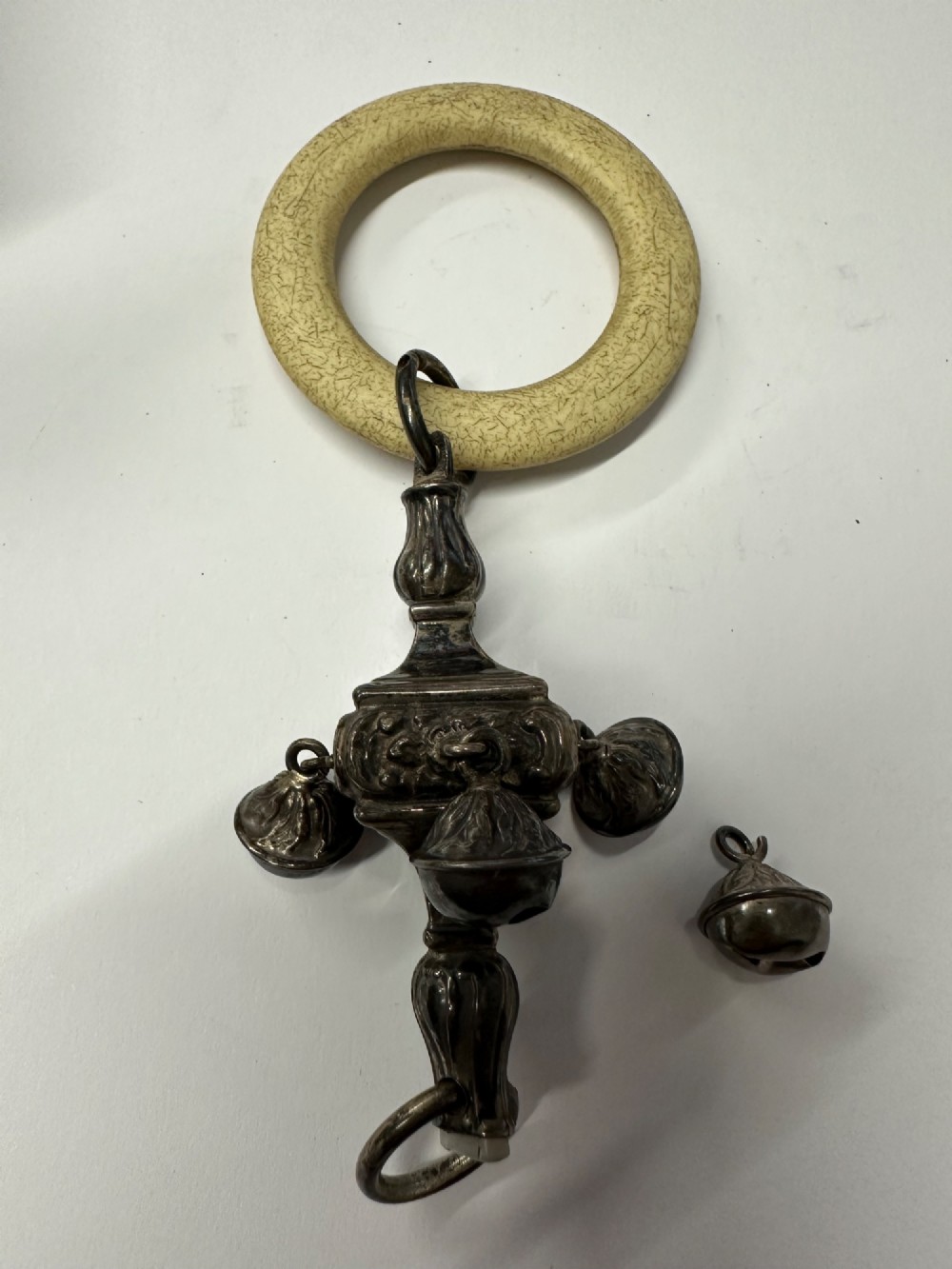 An antique silver baby rattle featuring embossed metalwork, small attached bells, and a cream-coloured circular teether ring. View XIII