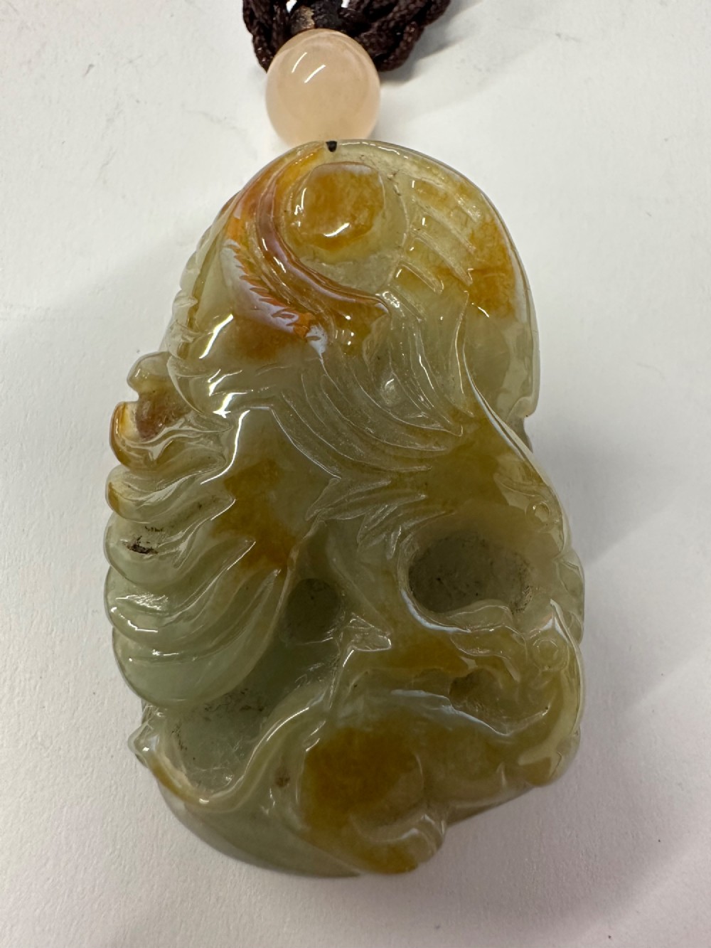 A hand-carved yellow and green jade pendant depicting a phoenix among foliage, suspended from a cord with a spherical accent bead.