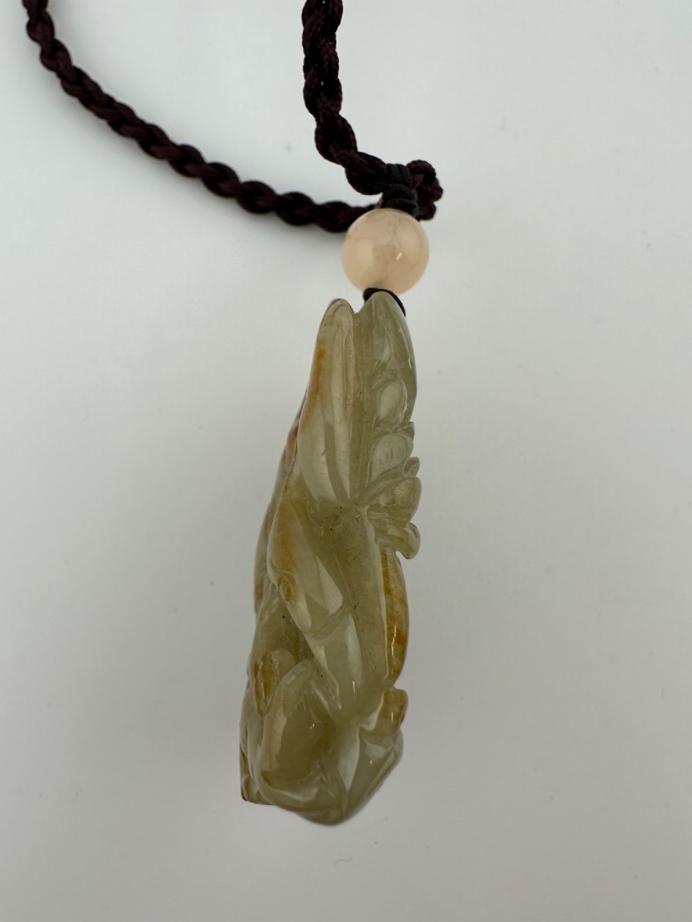 A hand-carved yellow and green jade pendant depicting a phoenix among foliage, suspended from a cord with a spherical accent bead. View VI