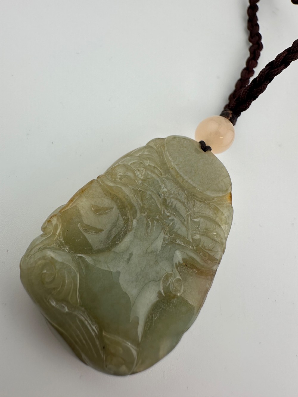 A hand-carved yellow and green jade pendant depicting a phoenix among foliage, suspended from a cord with a spherical accent bead. View X