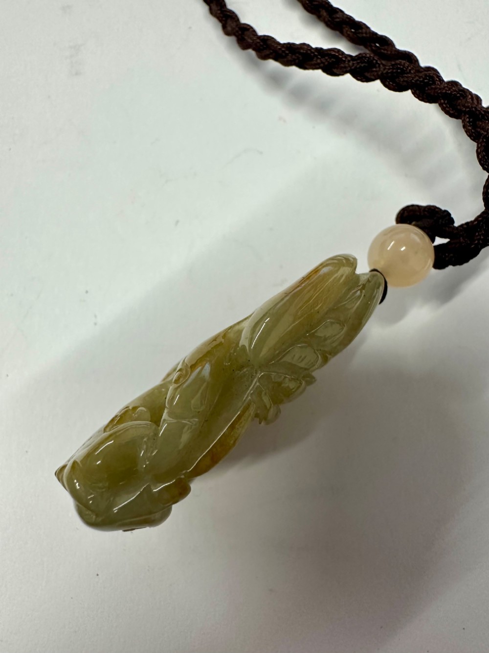A hand-carved yellow and green jade pendant depicting a phoenix among foliage, suspended from a cord with a spherical accent bead. View XIII