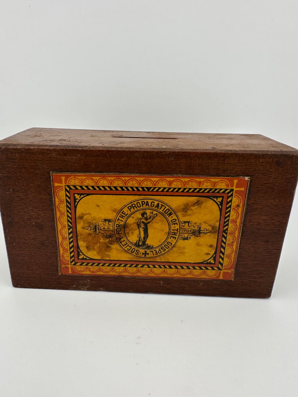 A rectangular dark wooden church collection box featuring a coin slot on top and a printed yellow and red Society for the Propagation of the Gospel label on the front.