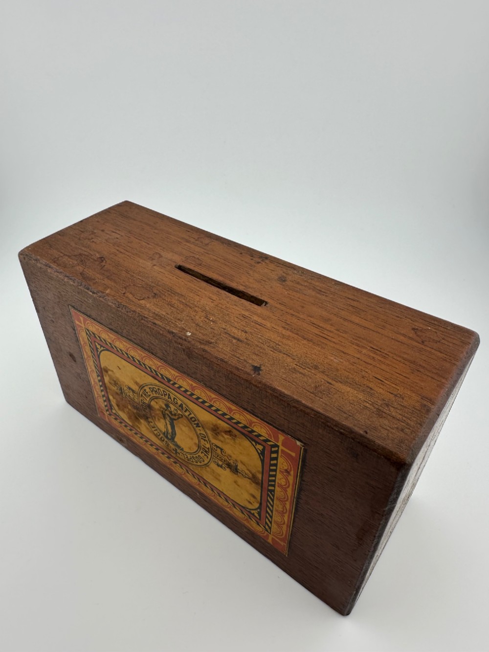 A rectangular dark wooden church collection box featuring a coin slot on top and a printed yellow and red Society for the Propagation of the Gospel label on the front. View II