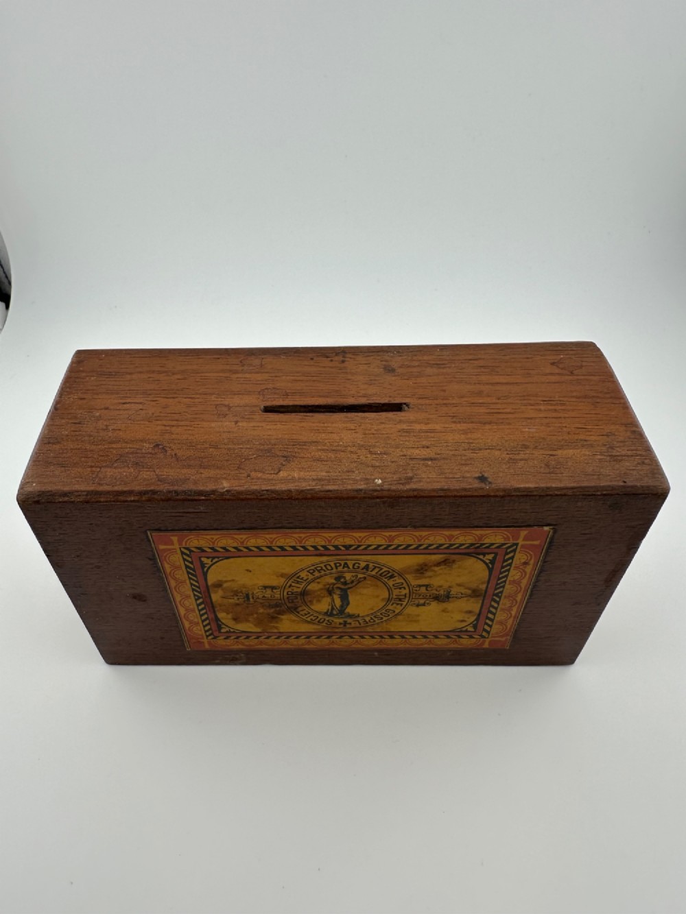 A rectangular dark wooden church collection box featuring a coin slot on top and a printed yellow and red Society for the Propagation of the Gospel label on the front. View IV