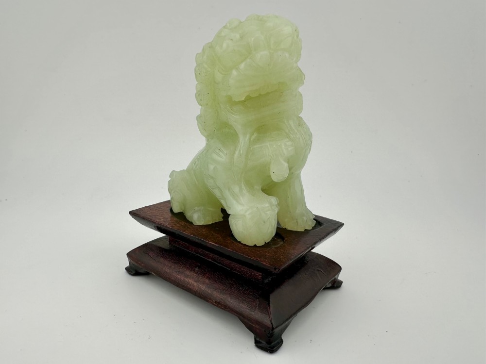 A hand-carved pale green jade guardian lion sculpture resting on a dark carved timber base.