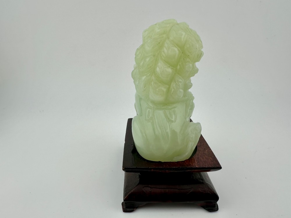 A hand-carved pale green jade guardian lion sculpture resting on a dark carved timber base. View III