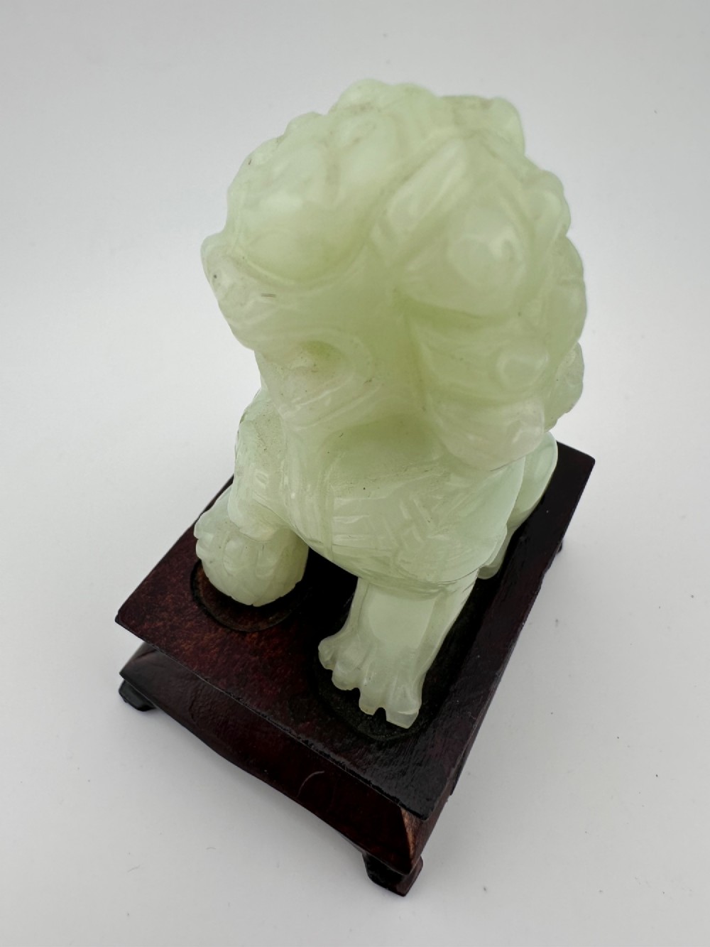 A hand-carved pale green jade guardian lion sculpture resting on a dark carved timber base. View V