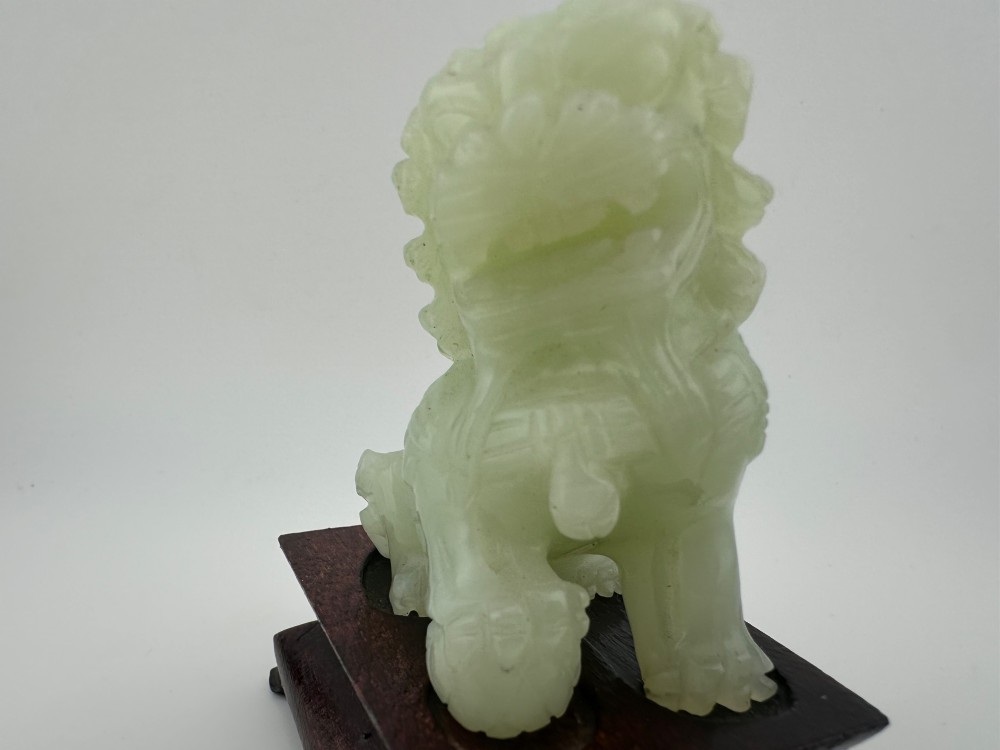 A hand-carved pale green jade guardian lion sculpture resting on a dark carved timber base. View VI
