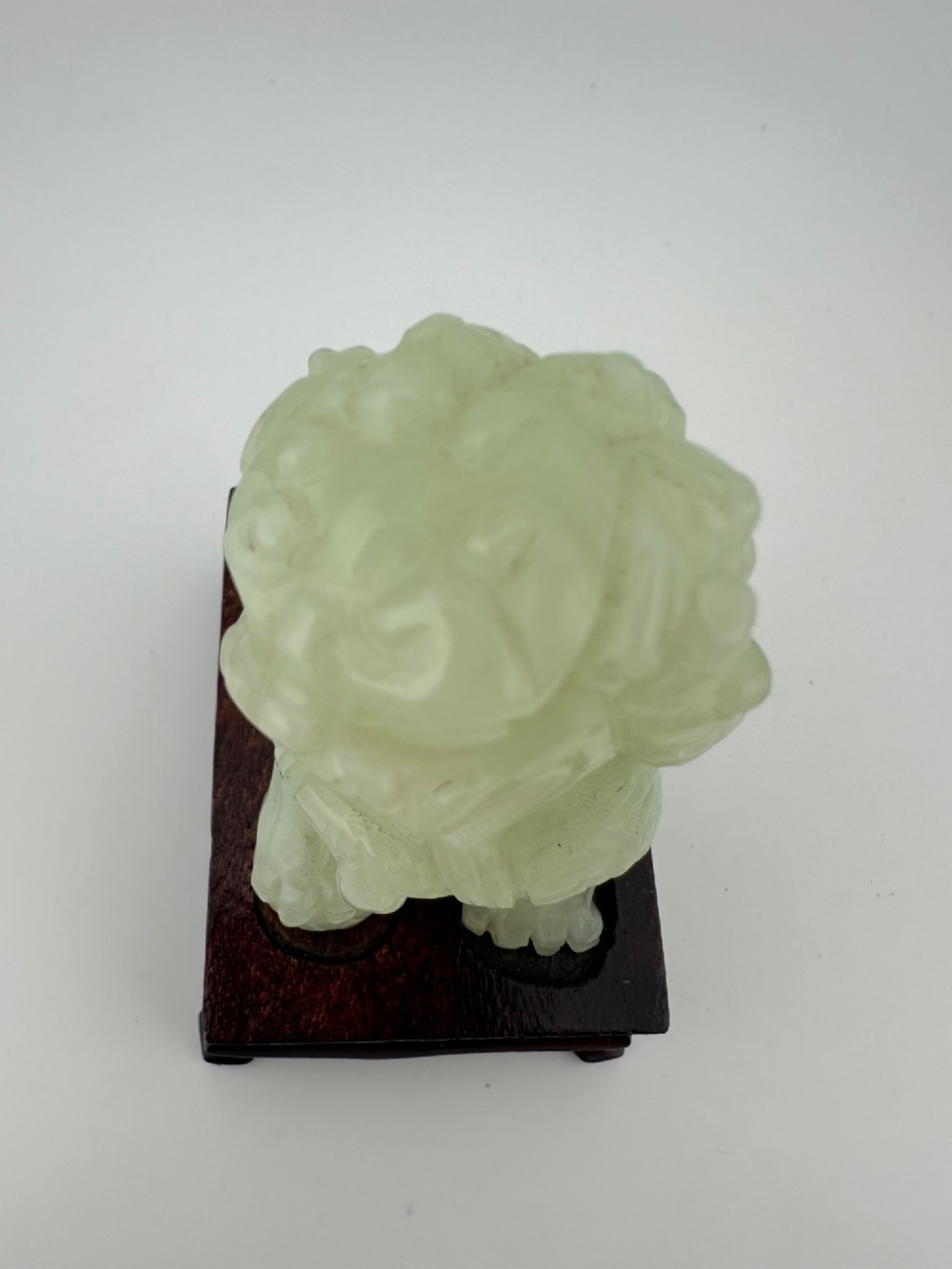 A hand-carved pale green jade guardian lion sculpture resting on a dark carved timber base. View VII