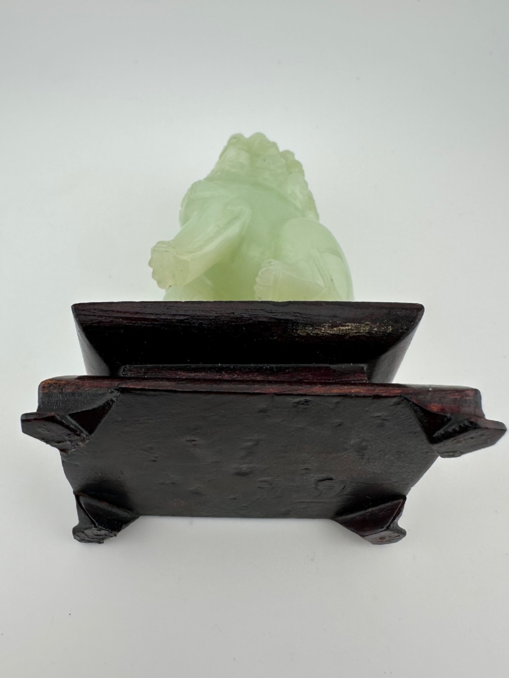 A hand-carved pale green jade guardian lion sculpture resting on a dark carved timber base. View X