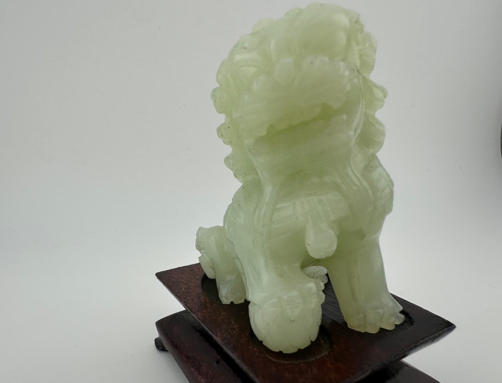 A hand-carved pale green jade guardian lion sculpture resting on a dark carved timber base. View XI