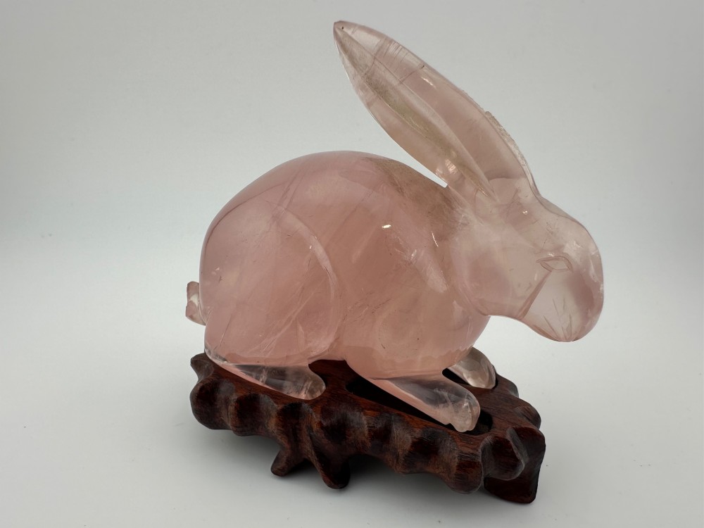 A hand-carved pink rose quartz rabbit figurine with upright ears seated on a dark fitted hardwood display base.