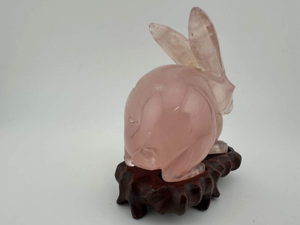 A hand-carved pink rose quartz rabbit figurine with upright ears seated on a dark fitted hardwood display base. View II