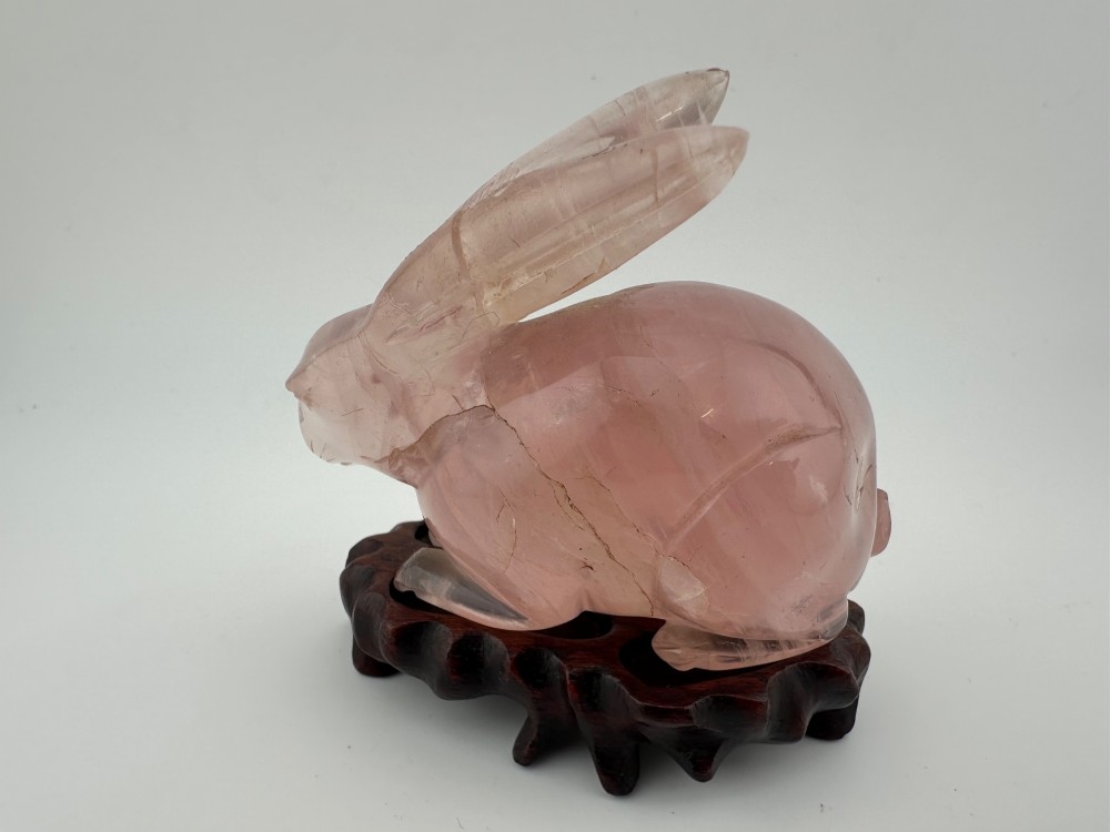 A hand-carved pink rose quartz rabbit figurine with upright ears seated on a dark fitted hardwood display base. View III