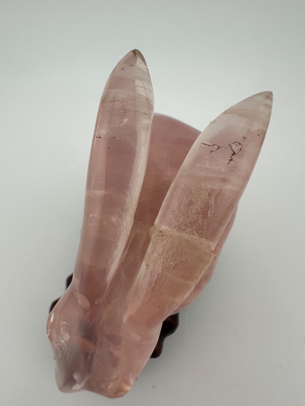 A hand-carved pink rose quartz rabbit figurine with upright ears seated on a dark fitted hardwood display base. View IV