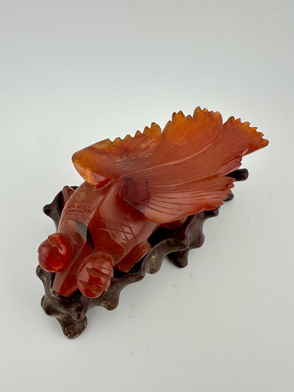 A translucent terracotta orange carnelian agate sculpture of a fantail goldfish positioned on a carved dark brown hardwood stand. View III