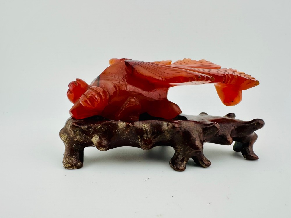 A translucent terracotta orange carnelian agate sculpture of a fantail goldfish positioned on a carved dark brown hardwood stand. View IX