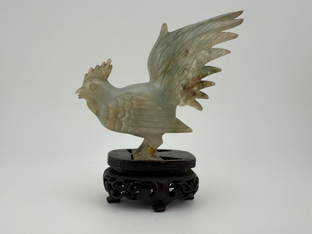 A hand-carved pale green jade sculpture of a cockerel with fan-shaped tail feathers, displayed on a dark carved hardwood base.