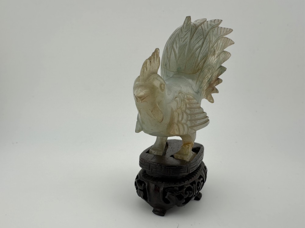 A hand-carved pale green jade sculpture of a cockerel with fan-shaped tail feathers, displayed on a dark carved hardwood base. View II