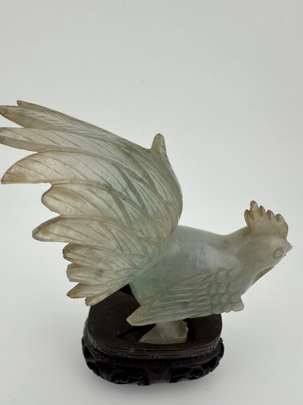 A hand-carved pale green jade sculpture of a cockerel with fan-shaped tail feathers, displayed on a dark carved hardwood base. View V