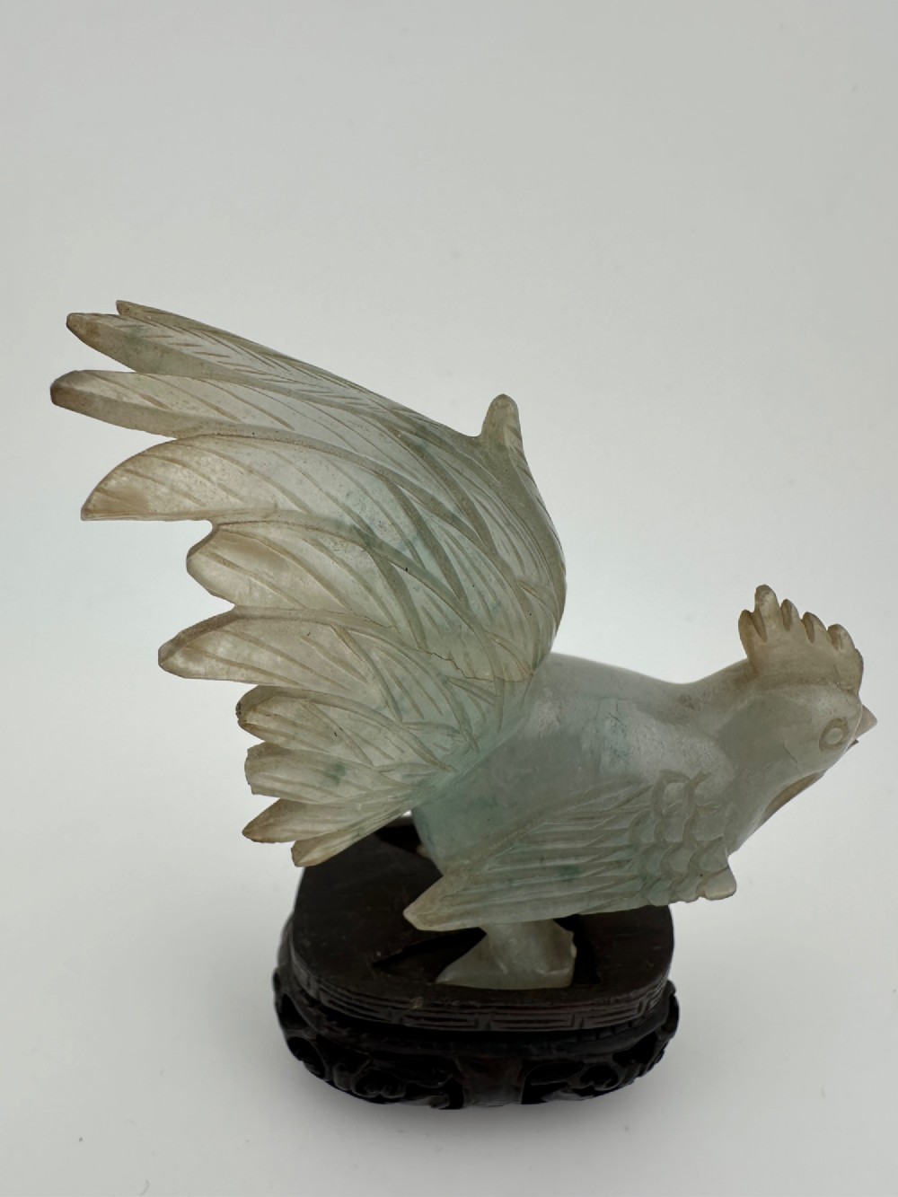 A hand-carved pale green jade sculpture of a cockerel with fan-shaped tail feathers, displayed on a dark carved hardwood base. View VI