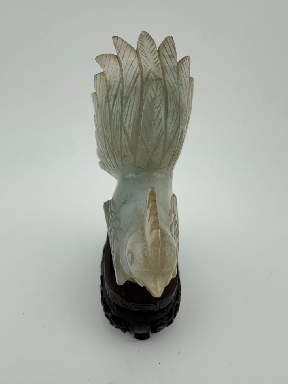 A hand-carved pale green jade sculpture of a cockerel with fan-shaped tail feathers, displayed on a dark carved hardwood base. View VII