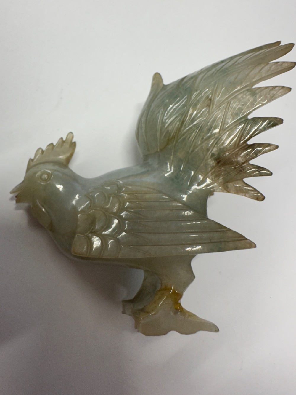 A hand-carved pale green jade sculpture of a cockerel with fan-shaped tail feathers, displayed on a dark carved hardwood base. View XIV