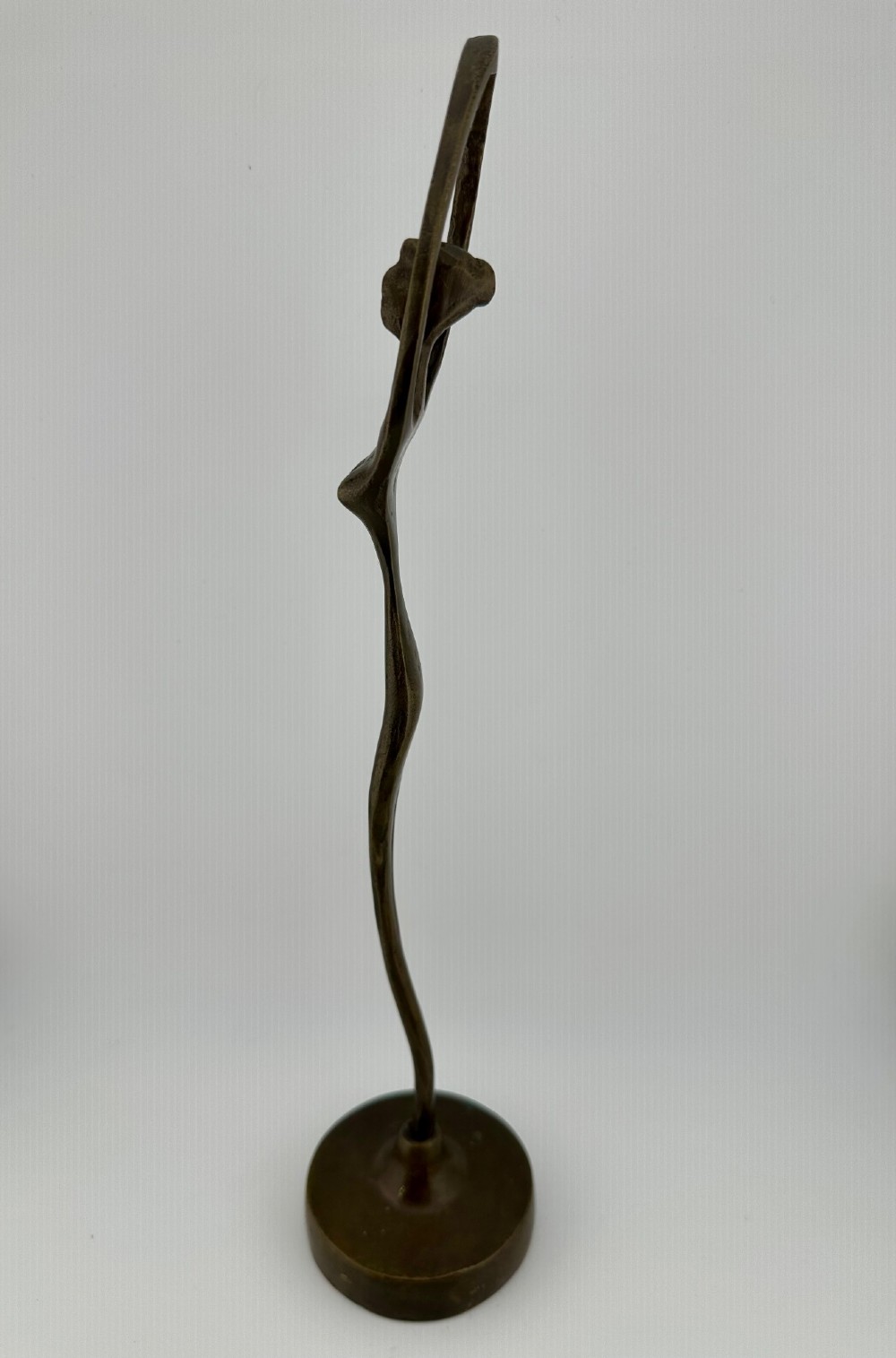 A slender modernist bronze sculpture depicting an abstract female dancer with raised arms set upon a round metal base. View III