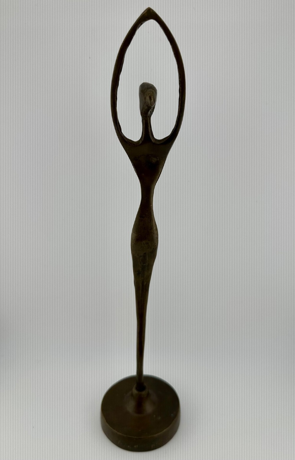 A slender modernist bronze sculpture depicting an abstract female dancer with raised arms set upon a round metal base. View IV