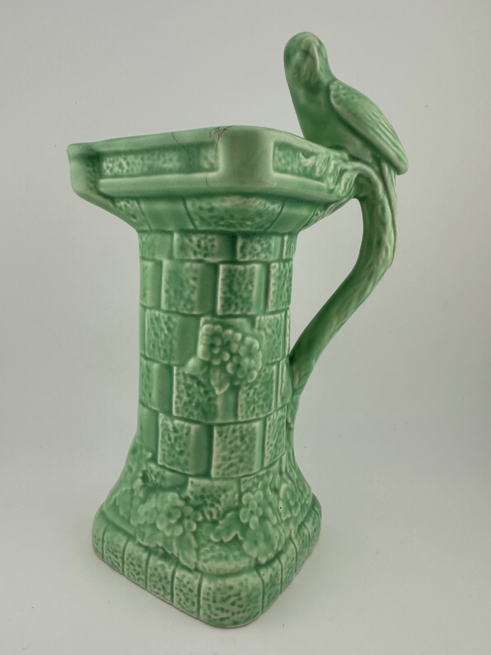 A pale green glazed earthenware Art Deco ceramic jug featuring relief brickwork and a sculpted bird perched on the handle.