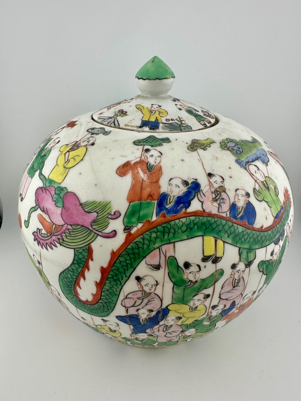 A rounded white porcelain covered jar featuring hand-painted colourful enamels depicting a dragon festival procession with children.