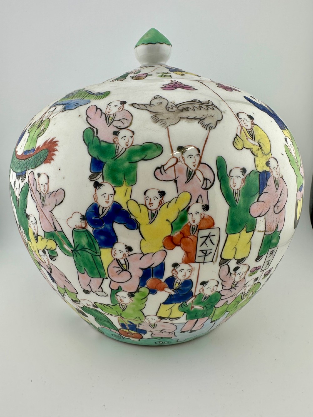 A rounded white porcelain covered jar featuring hand-painted colourful enamels depicting a dragon festival procession with children. View II