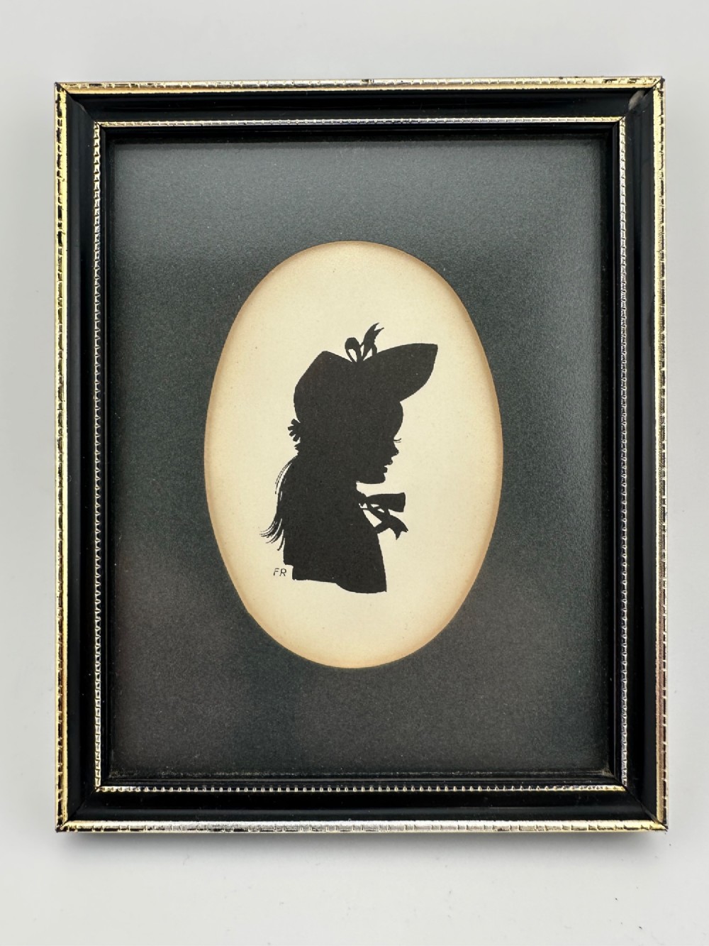A framed Art Deco silhouette portrait depicting a lady in profile with a decorative hat inside an oval cream mount surrounded by a black and gold frame.