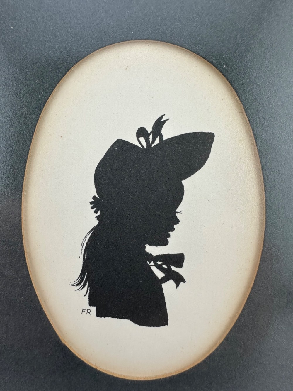 A framed Art Deco silhouette portrait depicting a lady in profile with a decorative hat inside an oval cream mount surrounded by a black and gold frame. View II